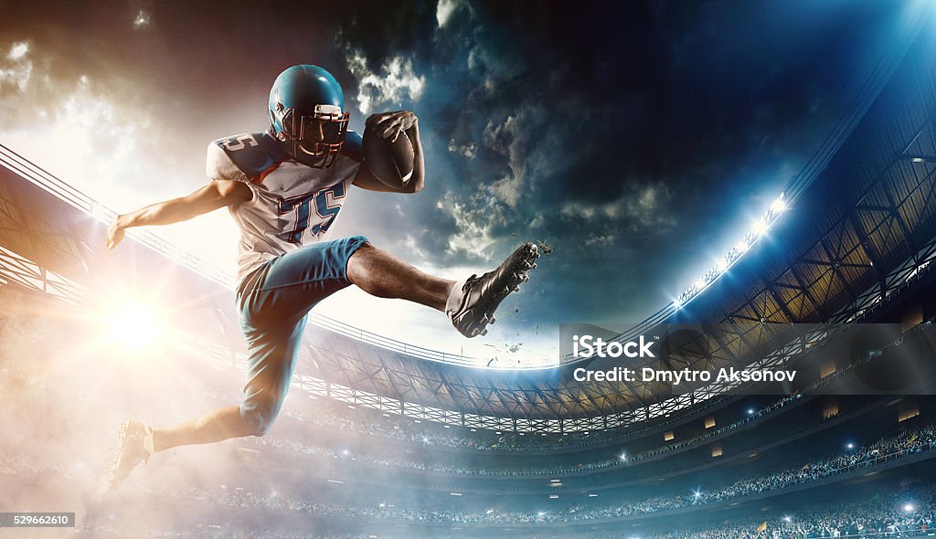 Football player runs with the ball Professional football player (Running Back) runs with the ball. The action takes place on professional stadium. The player wears unbranded sports uniform. There is artificial light on stadium together with sunlight. American Football - Sport Stock Photo