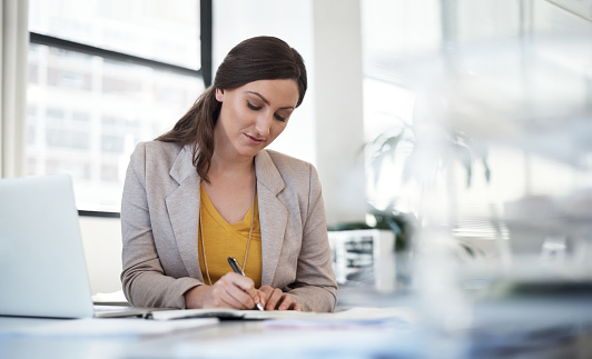 Shot of a young woman making notes at her desk in a modern office