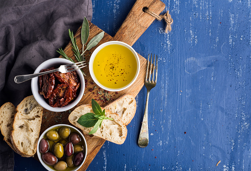 Mediterranean snacks set. Olives, oil, herbs and sliced ciabatta bread on yellow rustic oak board over painted dark blue background, top view, copy space