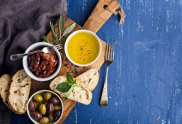 Mediterranean snacks set. Olives, oil, herbs and sliced ciabatta bread on yellow rustic oak board over painted dark blue background, top view, copy space