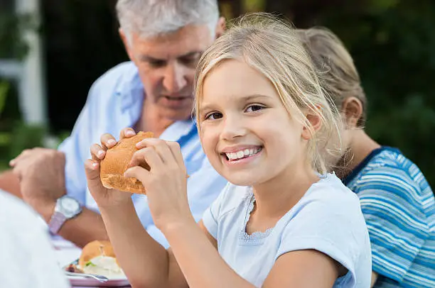 Young pretty girl enjoying eating bread and looking at camera. Little cute girl eating sandwich with brother and grandfather. Portrait of a young girl having lunch with family on picnic.