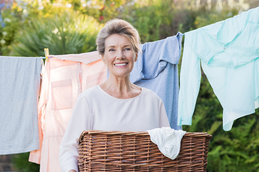Cheerful senior woman holding laundry basket in front of the clothesline. Portrait of old woman drying clothes. Senior woman hanging clothes out and looking at camera.