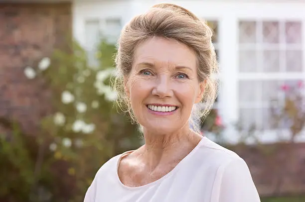 Happy senior woman standing outside her house. Content woman smiling and looking at camera. Portrait of cheerful old grandmother relaxing outside the house.