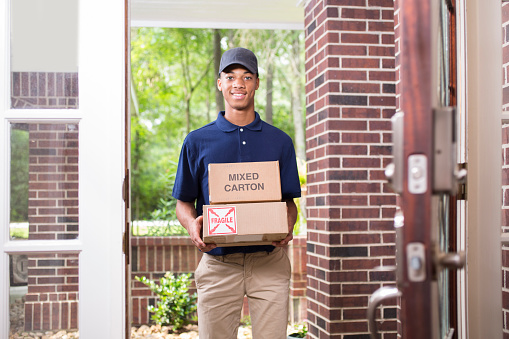 One young adult man wearing blue uniform delivers packages to customer at home setting.  The mixed race, African descent delivery person is holding two boxes and is looking at the camera as he gives the packages to you.   He stands in the customer's doorway, brick home entrance.