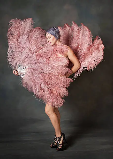 A burlesque dancer covers her body with feather fans and looks down.