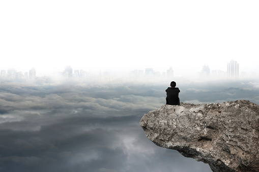 businessman thinking and sitting on cliff with gray cloudy sky cityscape background