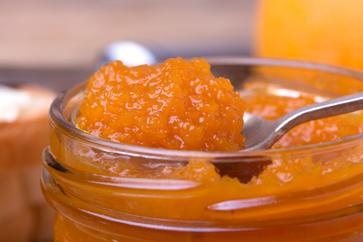 Closeup of colorful homemade orange jam in glass jar with spoon on rustic wooden board.