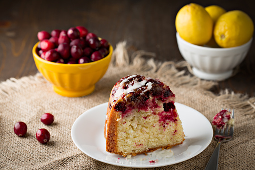 An extreme close up horizontal photograph of a small plate with a slice of a freshly baked holiday cranberry lemon bundt cake on a brown burlap placemat with a fork nearby and a white bowl and yellow bowls containing some ripe red cranberries  and tart lemons.
