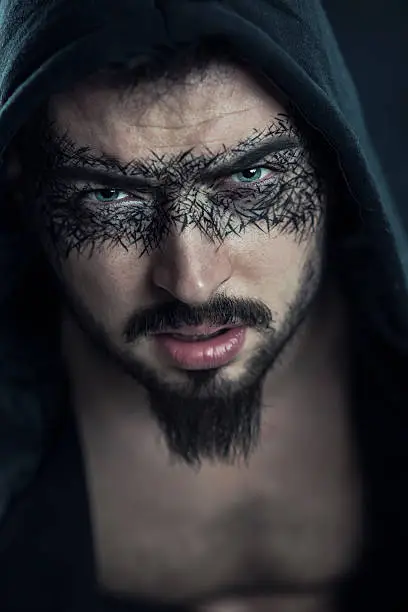 Young Man with beard and black make up around the eyes like superhero under the hood. Artistic Make-up and Head Scarf.