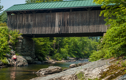 Belvidere, Vermont, USA- June 27, 2007: A teenager cools off on a hot summer day by using a rope swing to jump into a river below a covered bridge.