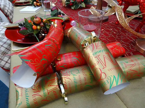 Photo showing a themed dinner table set for a Christmas meal featuring a decorative table cloth / runner, red and gold crackers, red tableware, crockery / plates, paper napkins, wine glasses and red pillar candles, with glittery centrepiece deer ornaments and golden ribbon decorations.