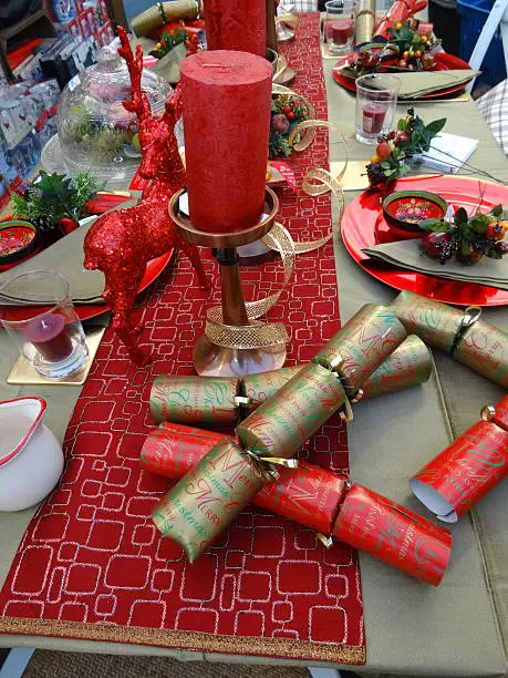 Photo showing a dinner table set for a Christmas meal featuring a decorative table cloth / runner, red and gold crackers, red tableware, crockery / plates, napkins, wine glasses and red pillar candles, with glittery centrepiece deer ornaments.