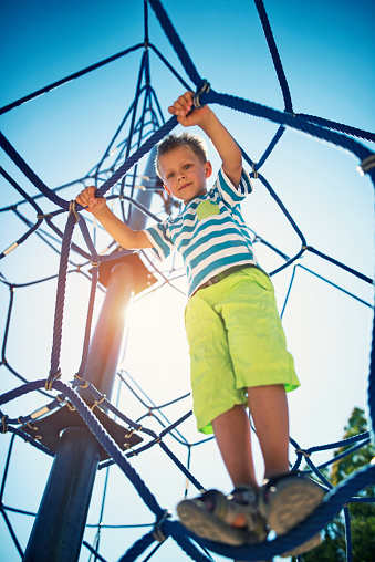 Portrait of little boy climbing in the playground. The boy aged 6 is looking down at the camera from the clibing web. The sun and blue sky visible in the background.