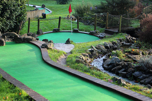 Photo showing a fun miniature golf course (adventure crazy golf), located in a public park.  The course layout features various hazards, synthetic putting greens with artificial felt grass and brick edging, log fences, rockeries, water features, bridges, garden areas and red flags in the holes.