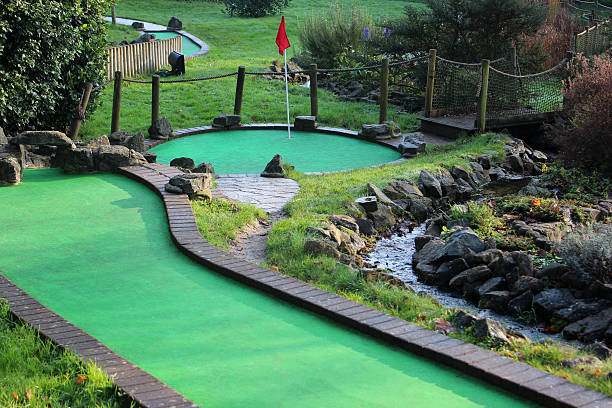 Image of miniature golf course (crazy golf), putting greens / flags Photo showing a fun miniature golf course (adventure crazy golf), located in a public park.  The course layout features various hazards, synthetic putting greens with artificial felt grass and brick edging, log fences, rockeries, water features, bridges, garden areas and red flags in the holes. putting green stock pictures, royalty-free photos & images