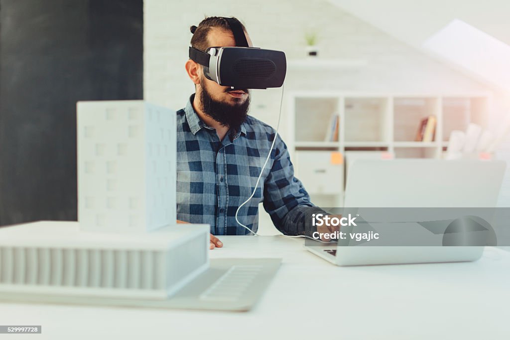 Architect using virtual reality glasses in his office. Modern Architect Using virtual reality glasses in his office. Virtually watching architecture model of business building. He is running virtual reality simulator on his laptop. *generic VR glasses Virtual Reality Simulator Stock Photo