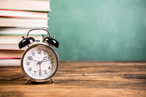 It's back to school time!  An alarm clock with stack of textbooks and an empty green chalkboard in background makes perfect copyspace for your message.  The group of objects lie on top of a wooden school desk.   Education background themes.