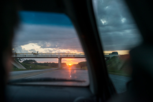 View from the backseat of a car traveling on a highway from Havana to Vinales, Cuba. The sun is setting, a horse-drawn cart crosses the bridge, and man stands on the side of the road waiting to hitch a ride.