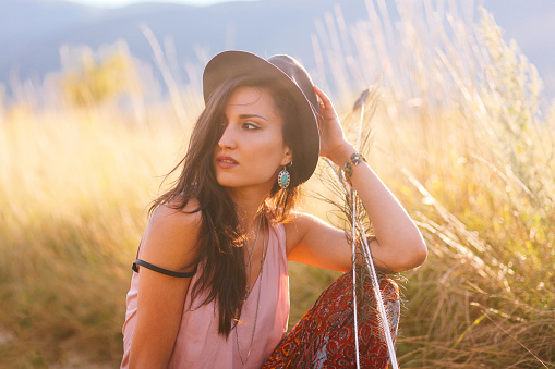 Vintage toned portrait of a young brunette in the beautiful outdoors just before sunset. She is wearing a hat and holding peackock feathers. 