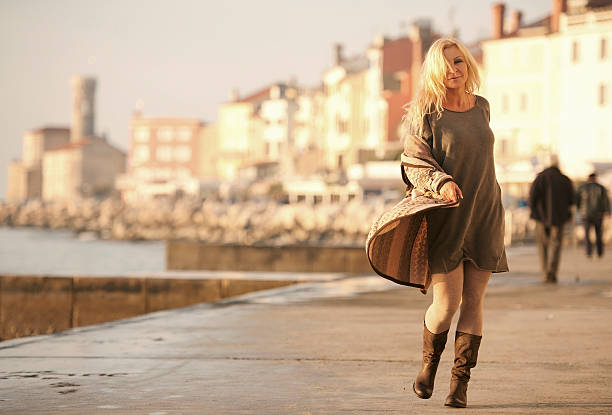 Attractive Mature Woman In Woolen Clothings Walking Attractive mature woman with long blond hair in woolen clothings in nature colors walking at seaside on a sunny winter day. Piran, Slovenia, Europe. older women short skirts stock pictures, royalty-free photos & images