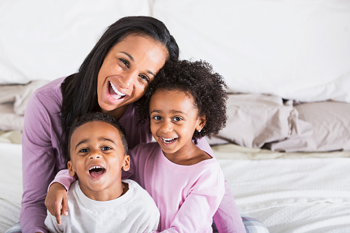 Portrait of an African American mother with twin 5 year old children, sitting on bed wearing pajamas.  They are smiling and laughing at the camera.