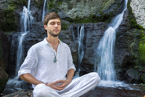 A young and handsome bearded man is doing yoga near a waterfall and profoundly meditates. Slow shutter speed photograph to smoothen the water falling from the rocks.