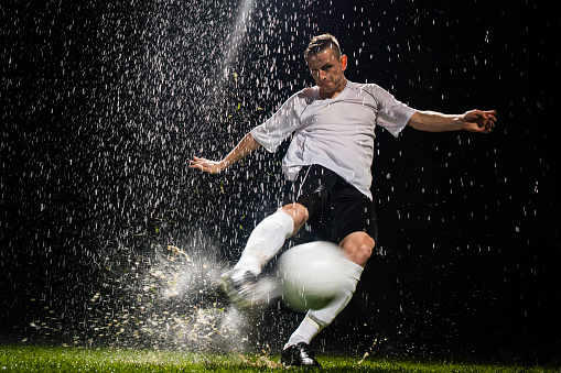 Soccer player kicks ball with full power in the rain at night.