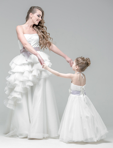 Beautiful blonde young woman dressed a wedding gown. Charming blonde little girl dressed a white dress. The bride and the little girl are dancing holding hands. The happy young woman and little girl are looking each other. Studio shooting on white background