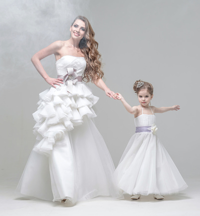 Beautiful blonde young woman dressed a wedding gown. Charming blonde little girl dressed a white dress. The bride and the little girl are dancing hand in hand. The happy young woman and little girl are looking at the camera. Studio shooting on white background