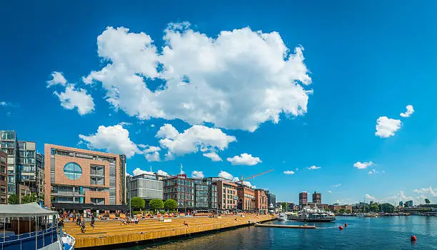 Big blue skies and white fluffy clouds over the sunlit boardwalks, cafes, restaurants and apartment buildings of the popular Aker Brygge district of central Oslo overlooking the tranquil harbour fjord, Norway. Composite panoramic image created from five contemporaneous sequential photographs. ProPhoto RGB profile for maximum color fidelity and gamut.