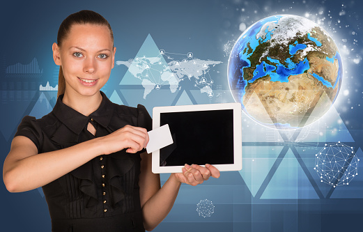 Beautiful businesswoman holding blank tablet PC and blank business card in front of PC screen. Globe, world map, network with people icons, triangles and other virtual elements as backdrop. Elements of this image furnished by NASA