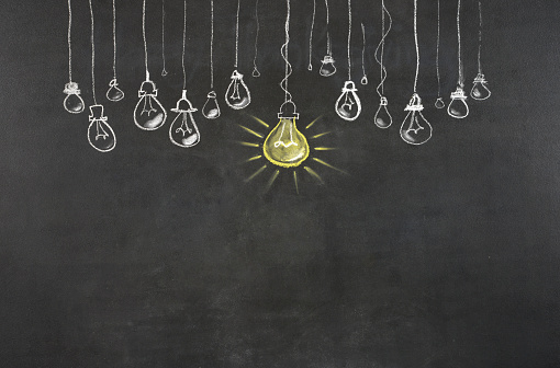 The chalkboard drawing shows multiple light bulbs in a row with one glowing. The light bulbs are hanging from the top. The small light bulbs are outlined in white,the big light bulb is yellow. The big light bulb is a symbol of innovation e/o success.