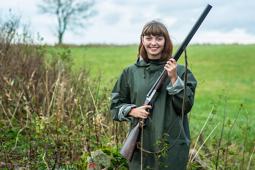 An attractive young woman outside on a field with a shotgun smiling to camera.