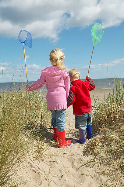 Little boy or girl wearing blue softshell overall walking on a sand dune with yellow shovel near the Baltic sea in autumn season