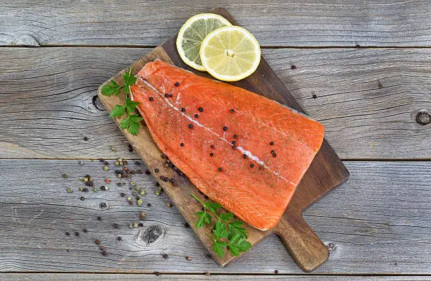 Top view image of a fresh salmon fillet with herbs, spices and lemon slices on rustic wood ready to be cooked.