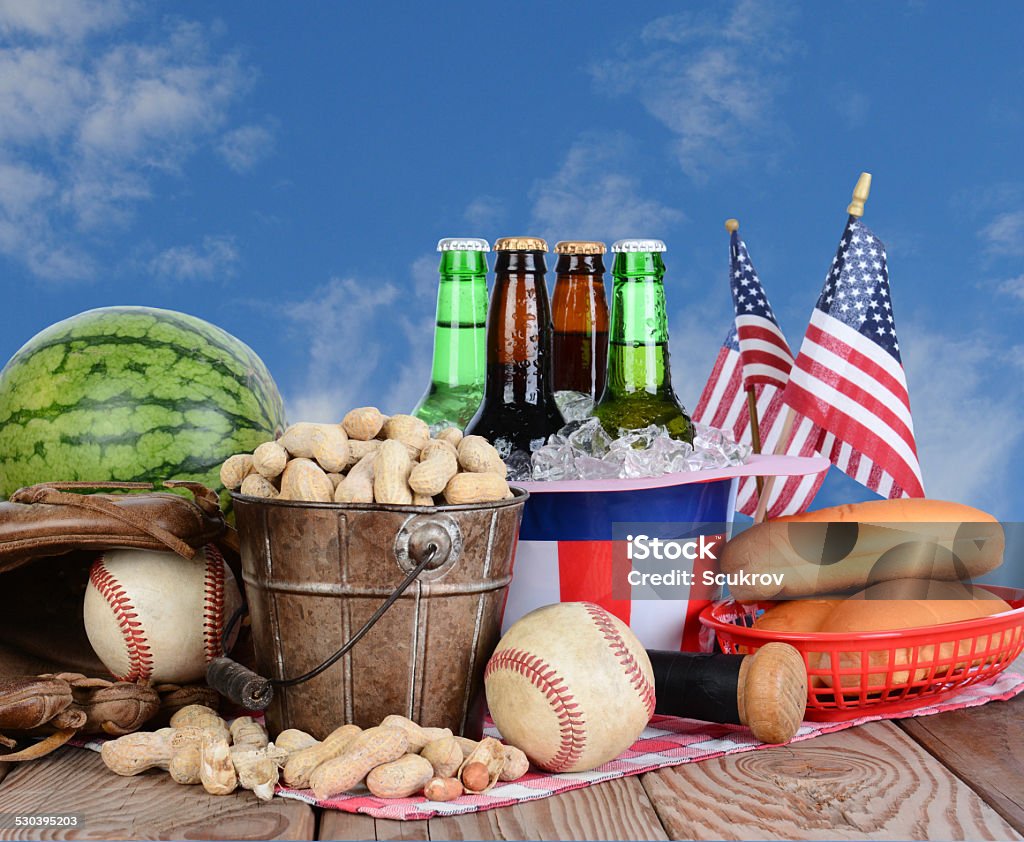 Fourth of July Picnic Table Picnic table ready for a Fourth of July celebration. Cold beer in an Uncle Sam Hat, watermelon peanuts, baseball, and hot dog buns fill the table, with a blue cloudy sky background. American Flag Stock Photo