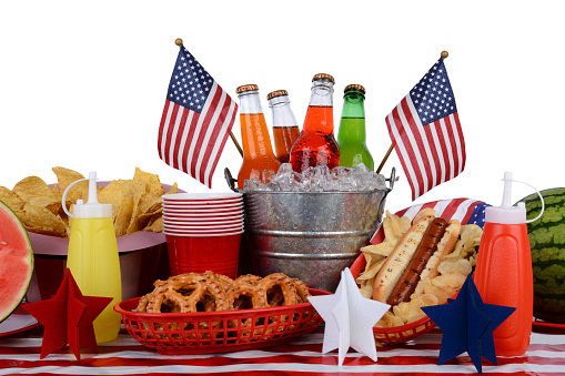 A picnic table set up with a Fourth of July theme. Horizontal format with a white background. Items include a soda bucket, hot dog, watermelon, chips, pretzels, and American Flag accessories.