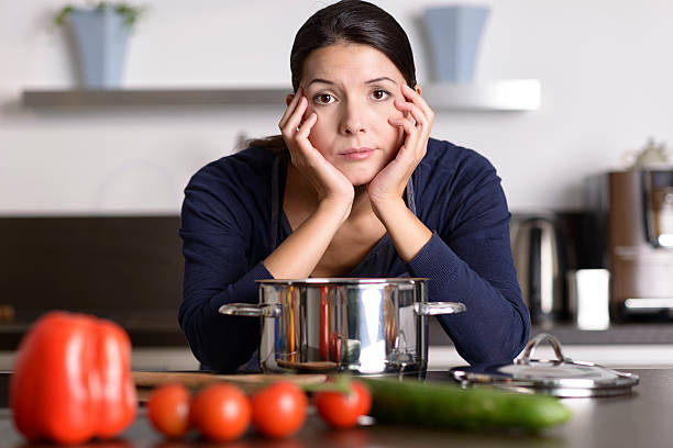 Unmotivated woman preparing the dinner Unmotivated attractive young woman preparing the dinner leaning on the hob eyeing the camera with a listless glum expression as she stands in her kitchen in an apron drudgery photos stock pictures, royalty-free photos & images