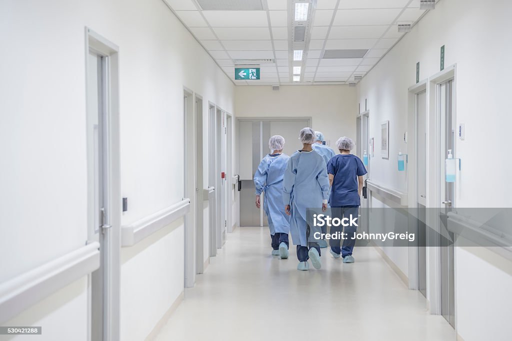 Rear view of surgeons walking down hospital corridor wearing scrubs View from behind of four doctors in hospital corridor walking away from camera. Medical team in modern hospital corridor wearing surgical scrubs Hospital Stock Photo