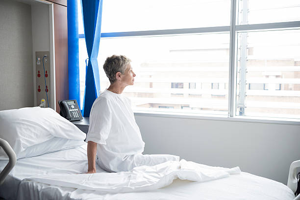 mujer mayor sentado sobre cama de hospital mirando a través de la ventana - examination gown fotografías e imágenes de stock