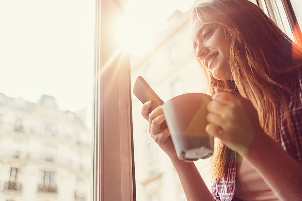 Happy woman with coffee cup texting on the open window Smiling woman using smartphone and drinking coffee in the morning house phone stock pictures, royalty-free photos & images