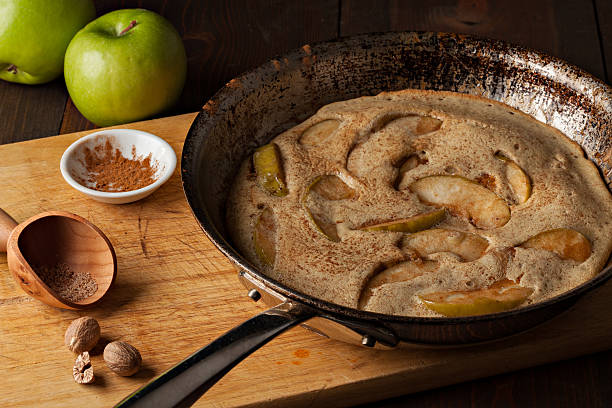 Dutch Baby Pancake A high angle extreme close up horizontal photograph  of a German apple pancake in grandmas well used frying pan. A bowl with ground cinnamon and a small wooden scoop along with ground nutmeg and some whole nutmegs sit by the pan on the cutting board.Two fresh green granny smith apple can be seen in the background. apple cinnamon pancake stock pictures, royalty-free photos & images