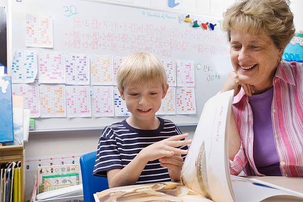 istock Boy Reading Book With Teacher In Classroom 530447357