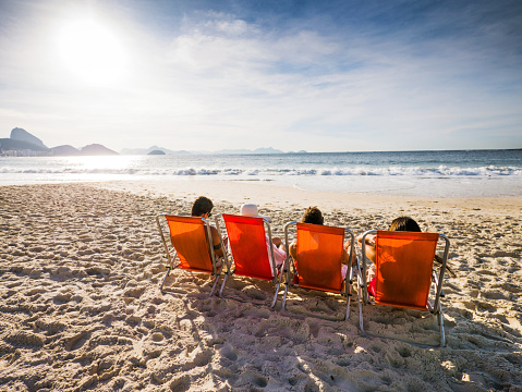 Rear view of four people sitting in deck chairs on the beach and enjoying the day.