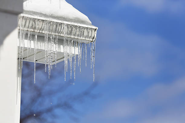 Icicles Melting Over Roof Edge Gutter A row of large frozen icicles is melting on a sunny winter day with a blue sky beyond. The icicles are hanging over the edge of an aluminum gutter on a residential home roof. icicles melting stock pictures, royalty-free photos & images