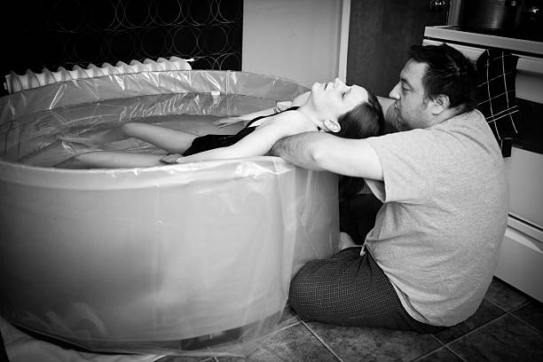Husband Comforts Wife During Home Water Birth Black and white photo of a husband comforting his wife during a water birth at home. water birth stock pictures, royalty-free photos & images