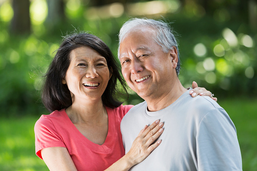 Portrait of a an affectionate senior Japanese couple standing outdoors in the park.  The woman has her arm wrapped around her husband and hand resting on his shoulder, with the other hand on his chest.  They are looking at the camera, smiling.  We see only their heads and shoulders.  The background is green grass,and bushes, out of focus.
