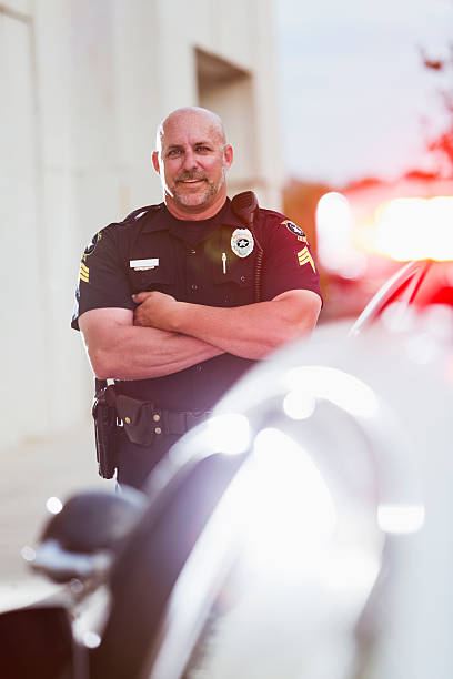 Happy police officer standing next to cruiser Policeman smiling, standing with arms crossed, looking at the camera. He is in his forties, with a bald head and mustache. A police car is out of focus in the foreground, with sirens flashing. police force smiling officer stock pictures, royalty-free photos & images