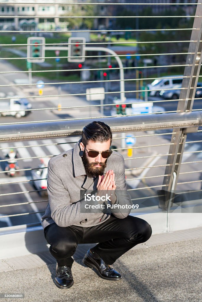 Young man (hipster style) sitting over a walk-bridge Young man (hispter style - well dressed with papillon) with big earring and full beard, wearing sunglasses sitting over a walk-bridge Hipster Culture Stock Photo