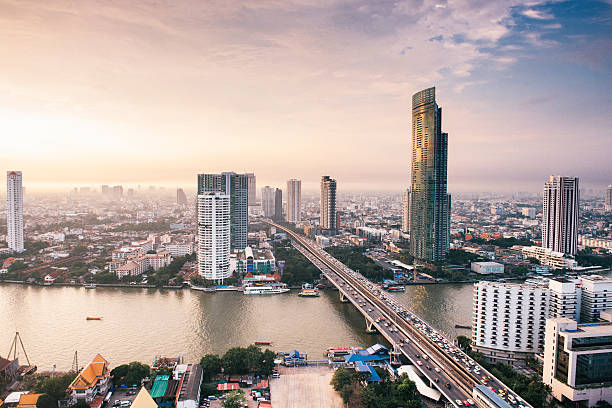 This is a horizontal, color photograph of Bangkok, Thailand. As the sun begins to set the sky turns colors above the high angle, scenic cityscape view seen from Sathorn Road. The Chao Phraya river is relatively empty. The bridge crossing the waterway is full of traffic. Photographed with a Nikon D800.